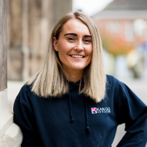 Amy Corfield is project manager at Kan Do Ventrues. Amy is photographed stood in a market square. The background is not in focus. Her light blonde hair is shoulder length and she is wearing a navy sweatshirt. She is smiling broadly.