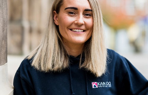 Amy Corfield is project manager at Kan Do Ventrues. Amy is photographed stood in a market square. The background is not in focus. Her light blonde hair is shoulder length and she is wearing a navy sweatshirt. She is smiling broadly.
